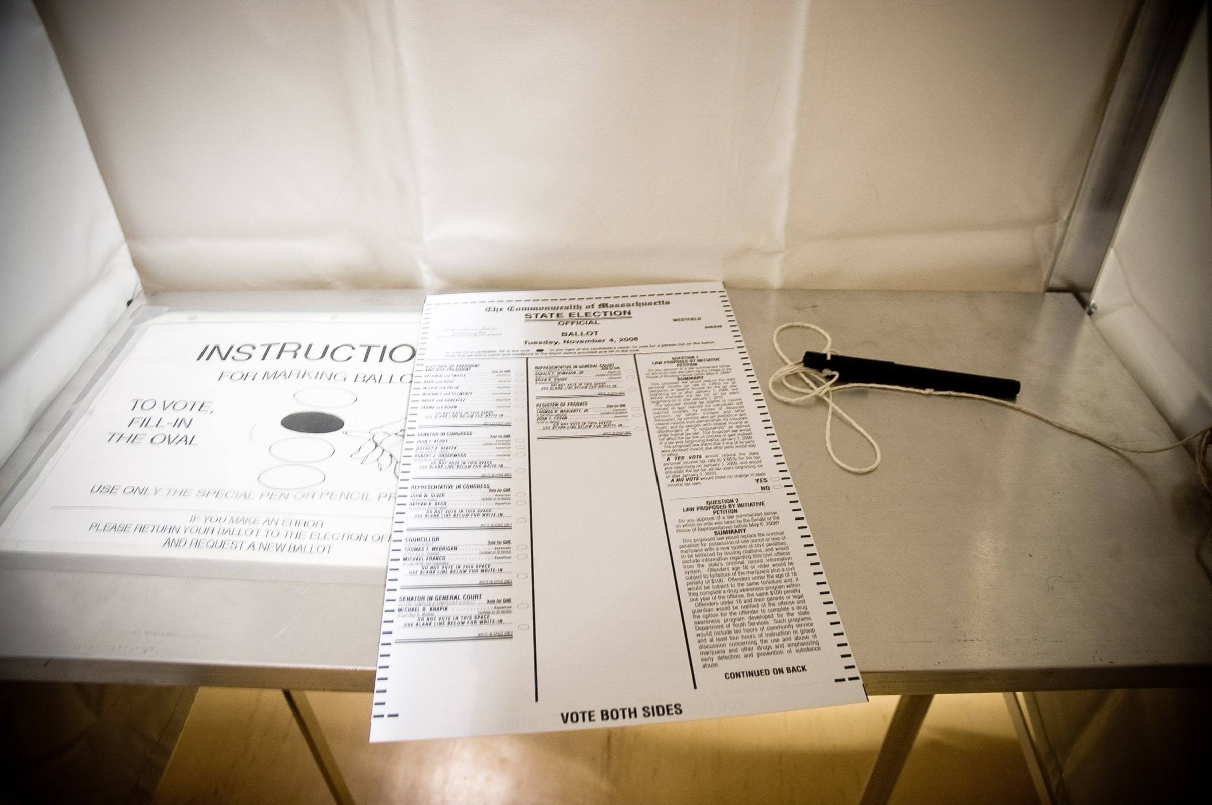 A paper election ballot resting on the metal shelf of a white voting booth, with a marking-instructions placard on the left panel and a pen tethered on a string to the right.