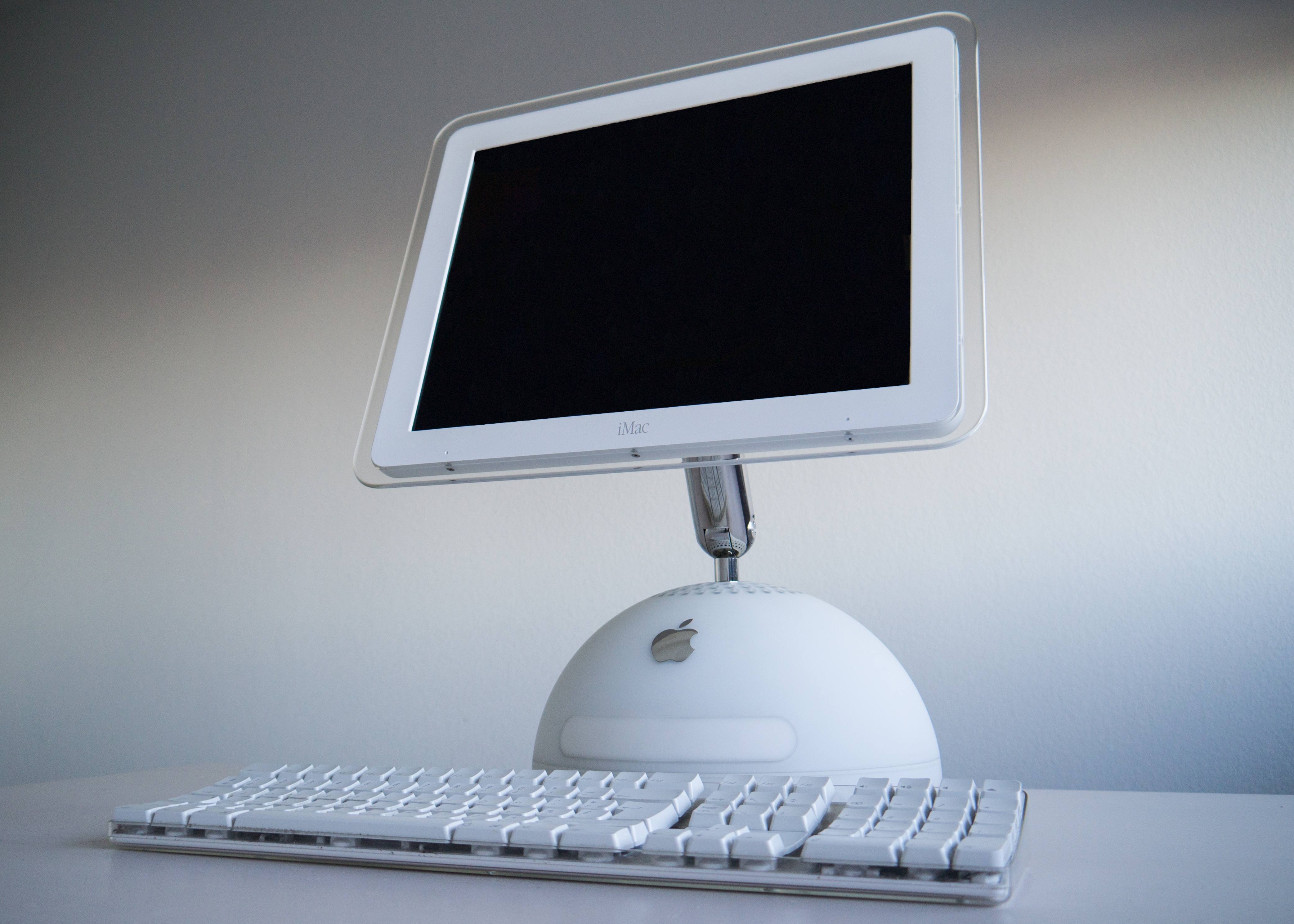 A white iMac G4 from 2002: a flat screen on a chrome arm above a white dome base, with a white keyboard in front.