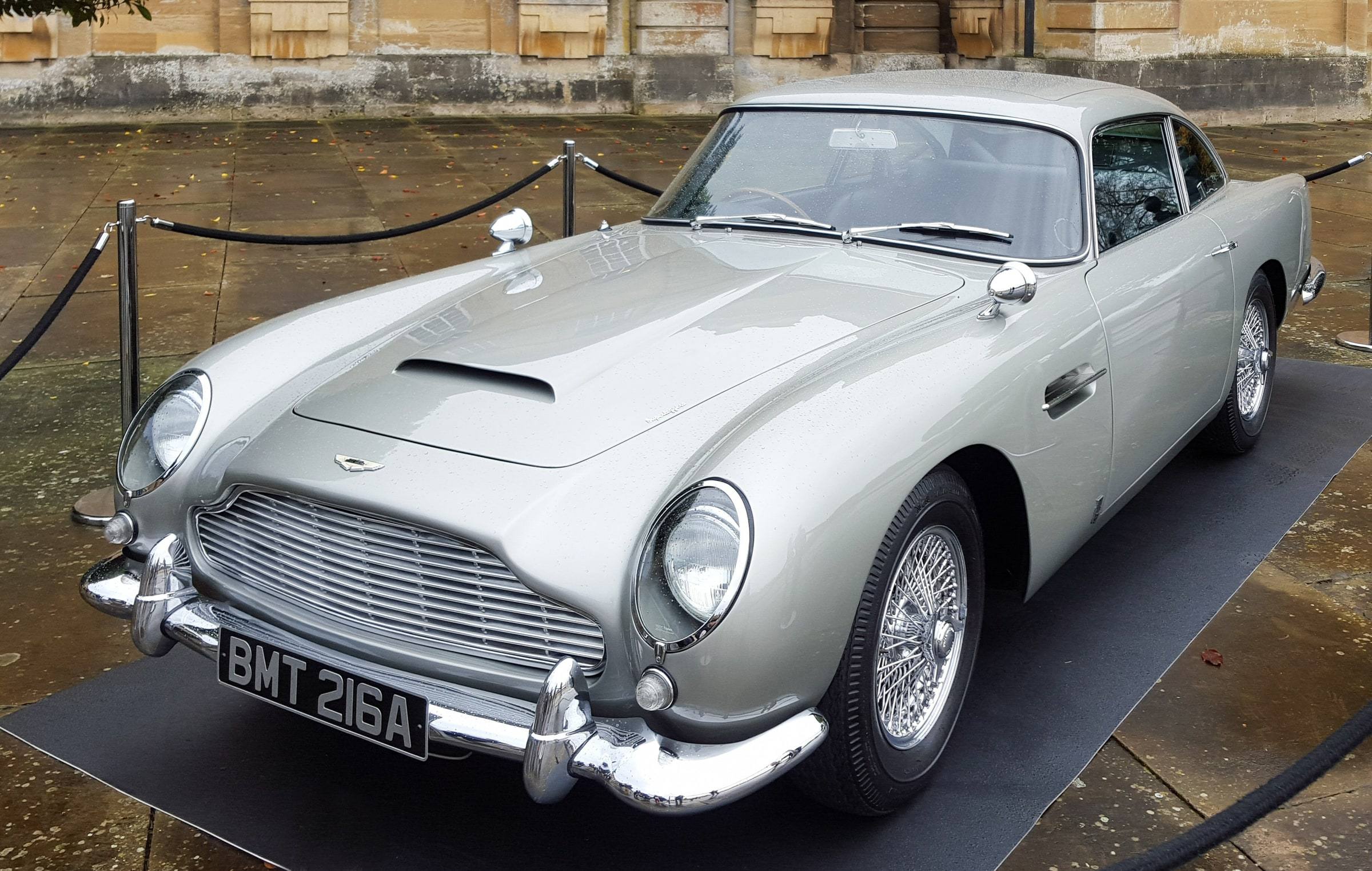 A silver Aston Martin DB5 with chrome bumpers, wire wheels and the number plate BMT 216A, parked on a black mat behind a velvet rope on a wet stone courtyard.