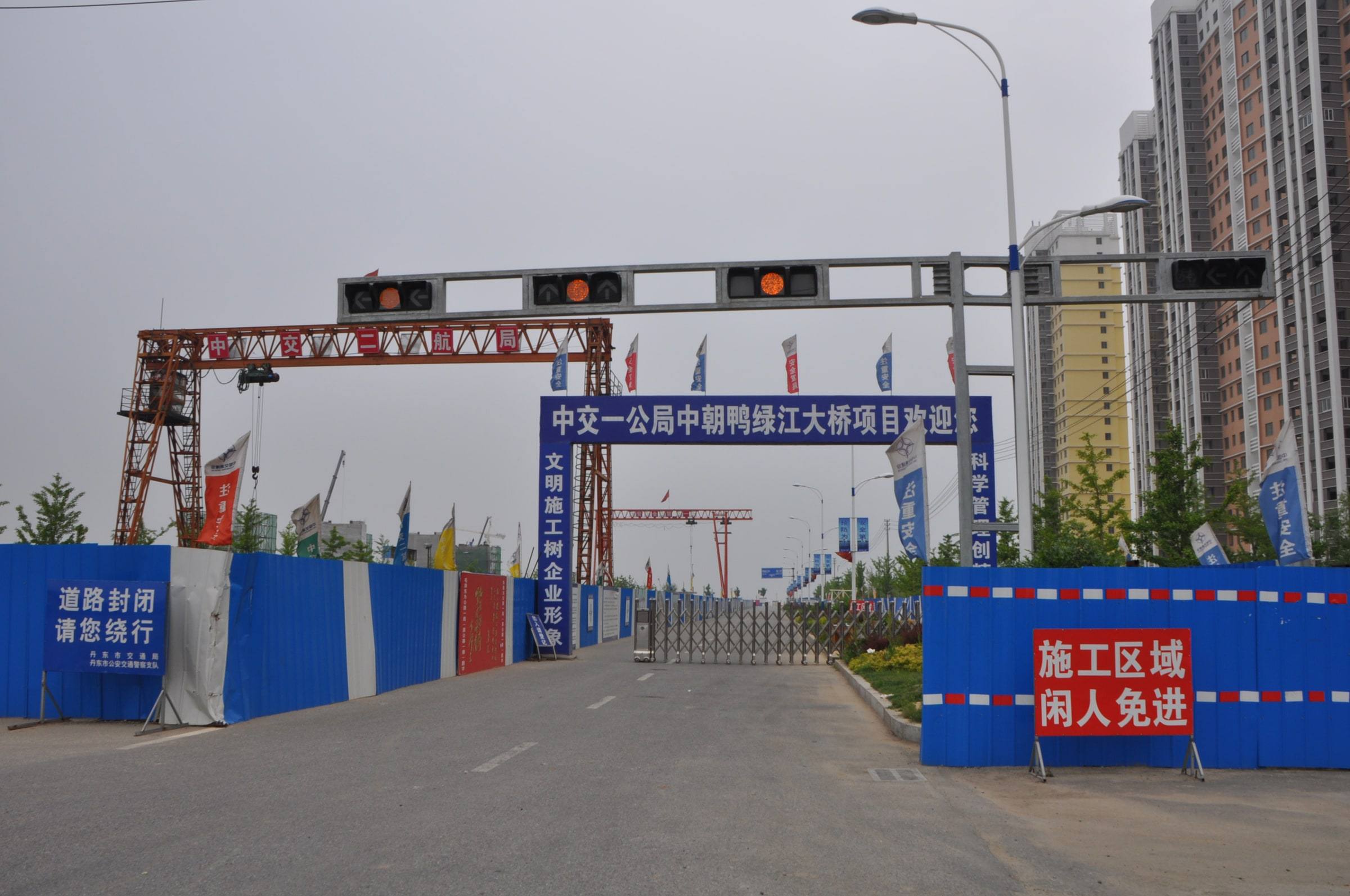 A closed road leads to a gated construction site under a blue banner and a gantry crane, with traffic lights overhead, blue barrier walls and a red sign in Chinese at right.