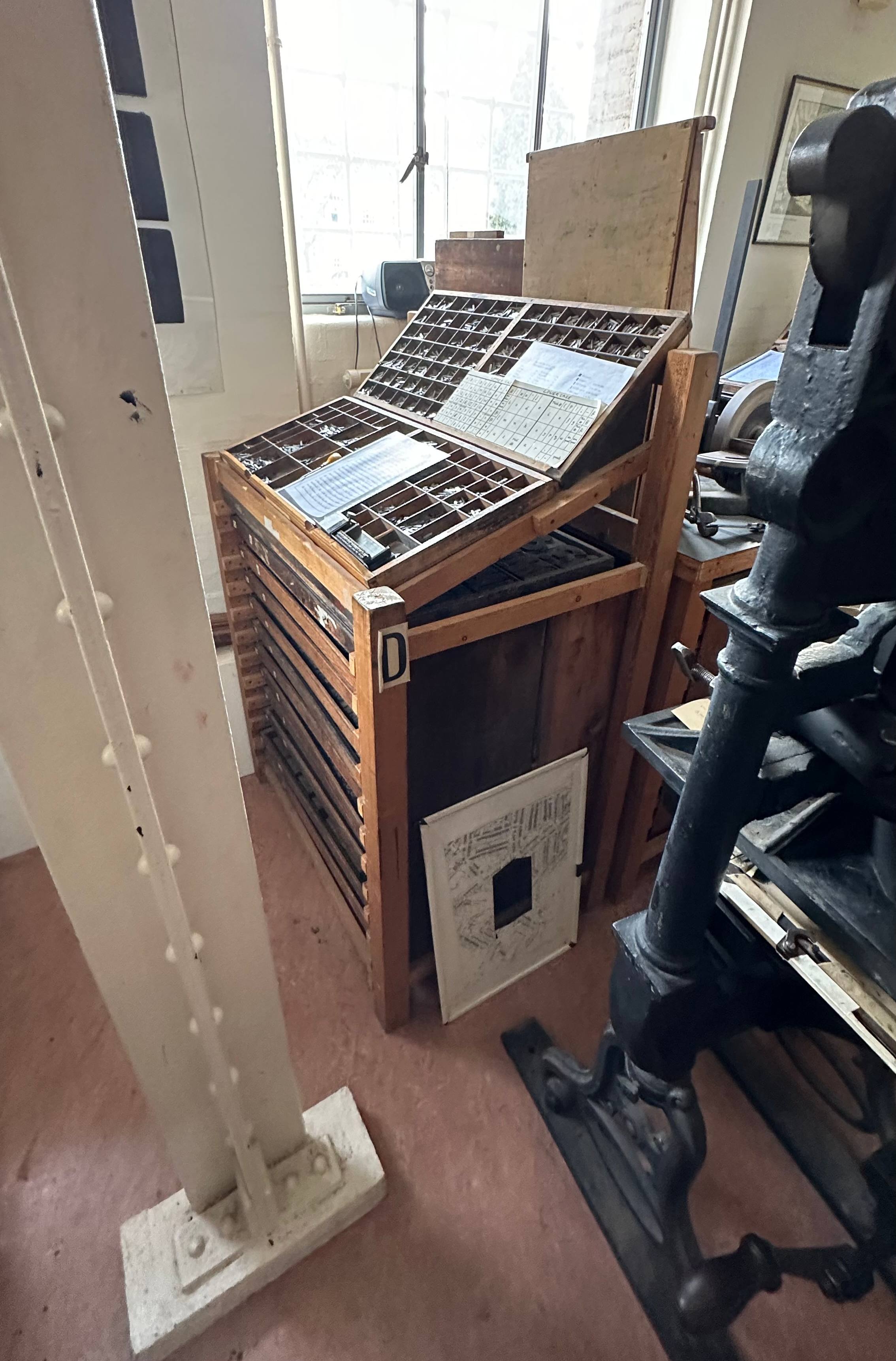 A quiet historical printing room: an old wooden cabinet of type cases with drawers of metal letters and a printed proof resting on top, beside the black iron frame of a hand press, under a tall window.