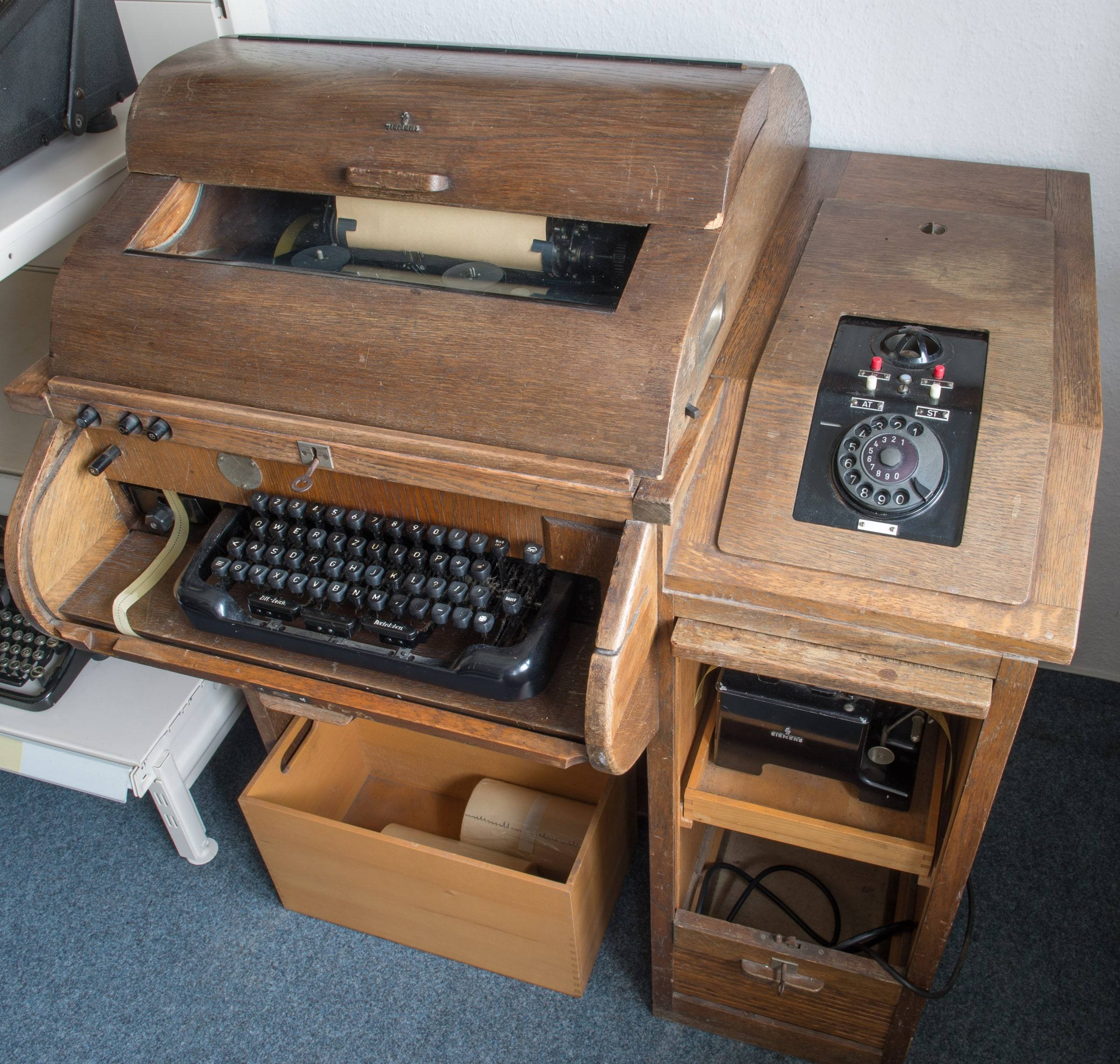 An old wooden Siemens teletypewriter with a roll-top cover raised over a paper feed, a black typewriter keyboard on a pull-out shelf, and a rotary telephone dial mounted on the right side, standing on a grey carpet in a museum.