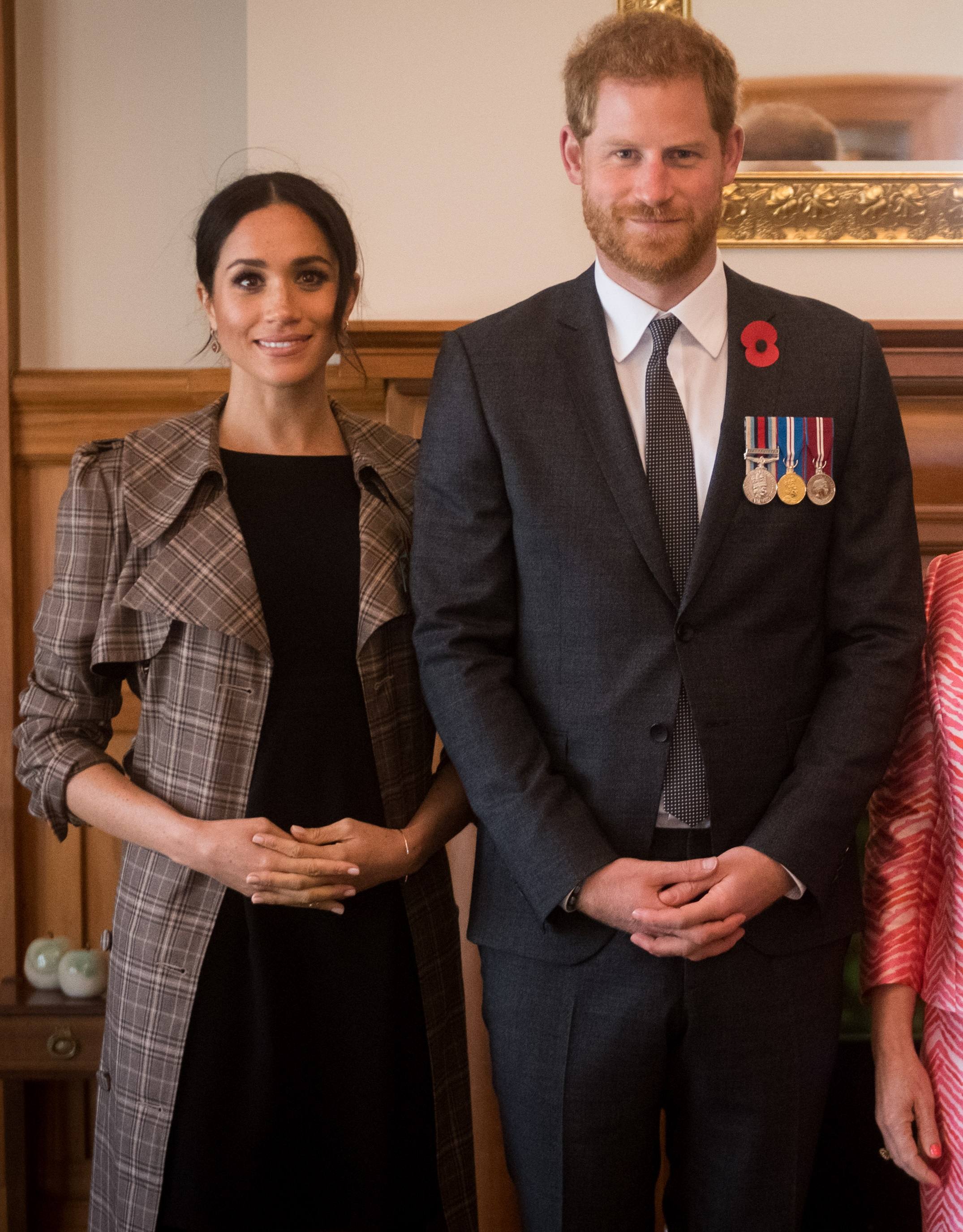 Meghan, in a plaid coat over a black dress, and Harry, in a dark suit with a red poppy and three medals, stand side by side smiling in a wood-paneled room.