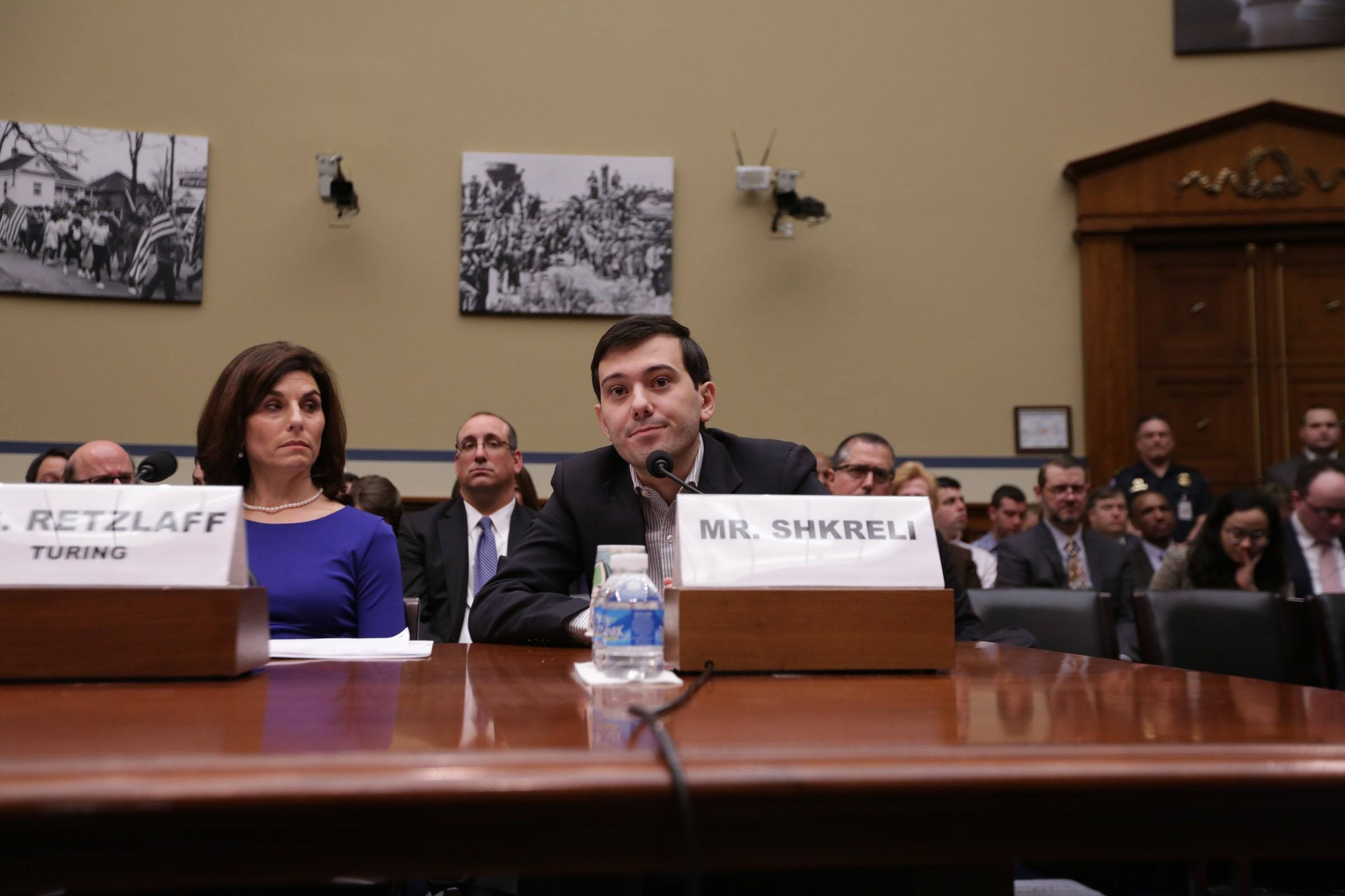 Martin Shkreli seated at a congressional witness table behind a microphone and a name card reading Mr. Shkreli, with a woman seated beside him and an audience behind.