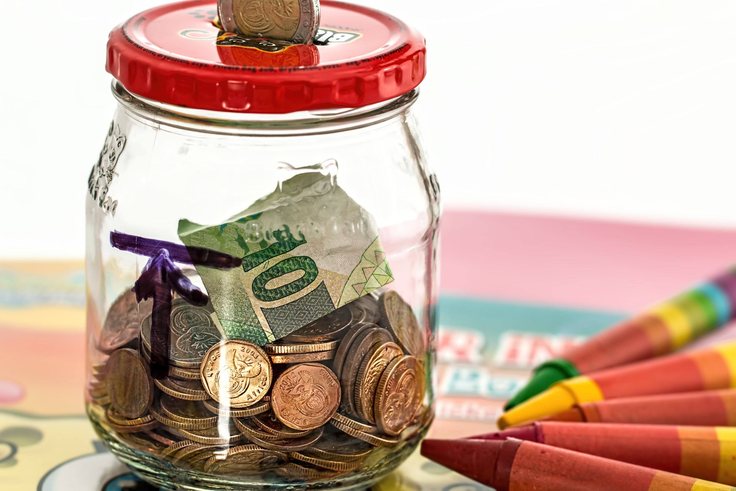 A clear glass savings jar with a red screw lid, a coin being dropped through the slot in the top, filled with assorted coins and a folded ten banknote, sitting on a colorful surface beside a row of children's crayons.