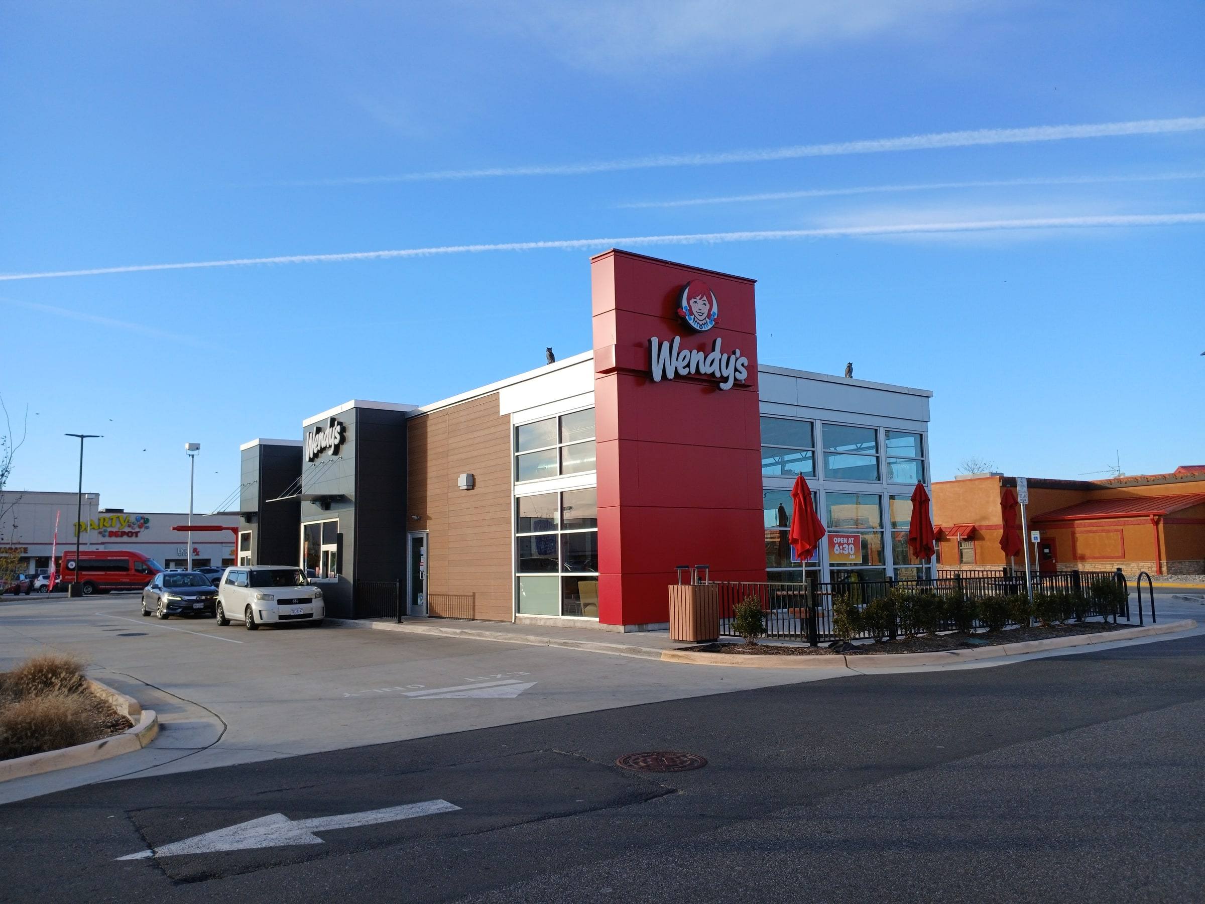 A modern Wendy's restaurant with a red and tan facade and the lit Wendy's logo on its tower, under a clear blue sky, with two cars in an otherwise empty parking lot out front.