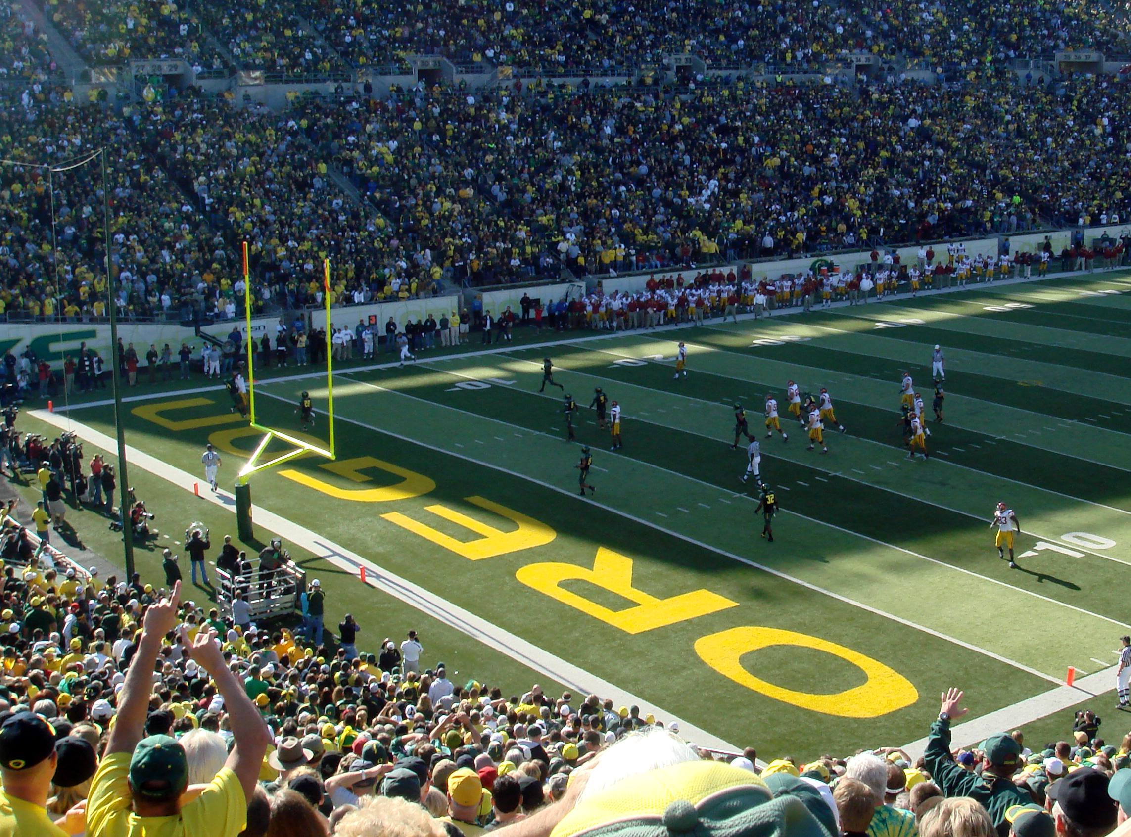 A college football game in a packed stadium, with players on the field near an end zone painted OREGON in yellow and a crowd of fans cheering in the stands.