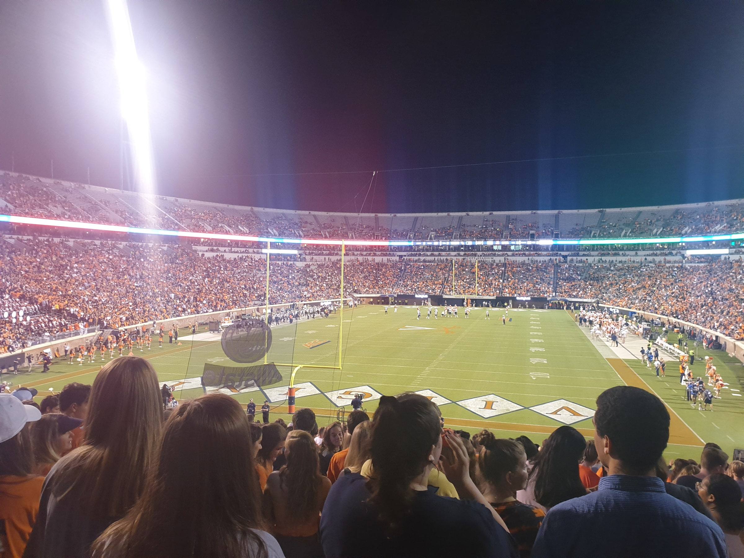 A packed stadium at night seen from the stands: a crowd of spectators in the foreground, the brightly lit green field and a yellow goalpost below, players on the field, and floodlights blazing against a dark sky.