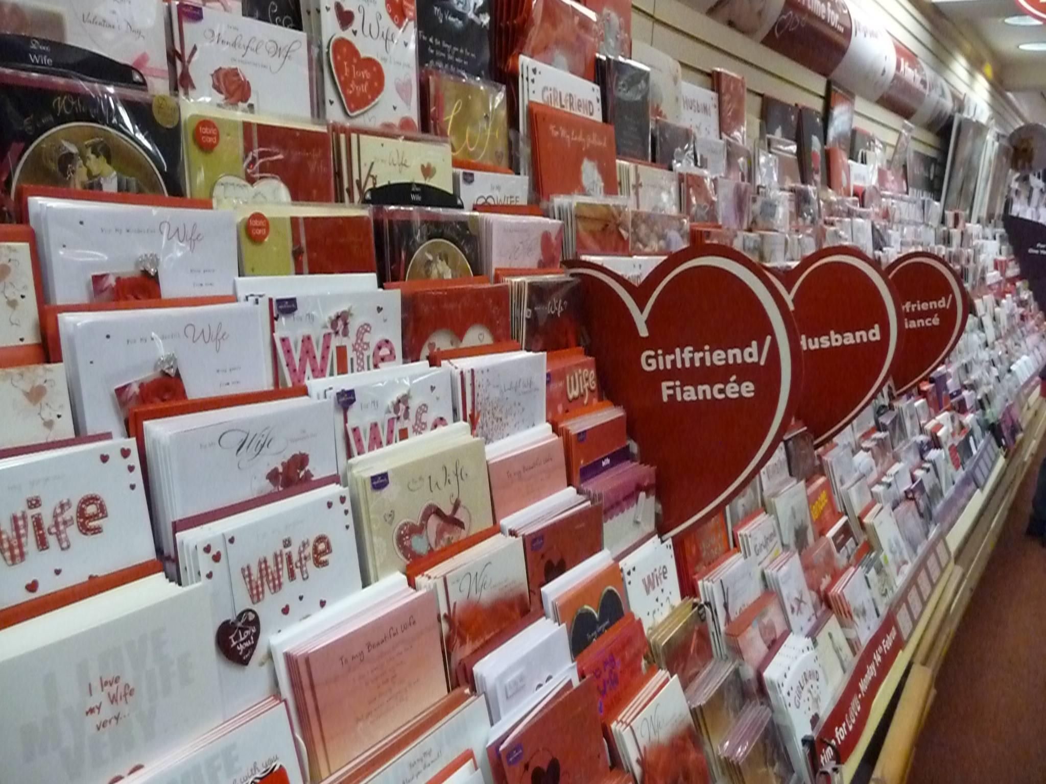 A long card-shop aisle packed with red and white love cards, sorted under heart-shaped signs reading Girlfriend/Fiancée and Husband.