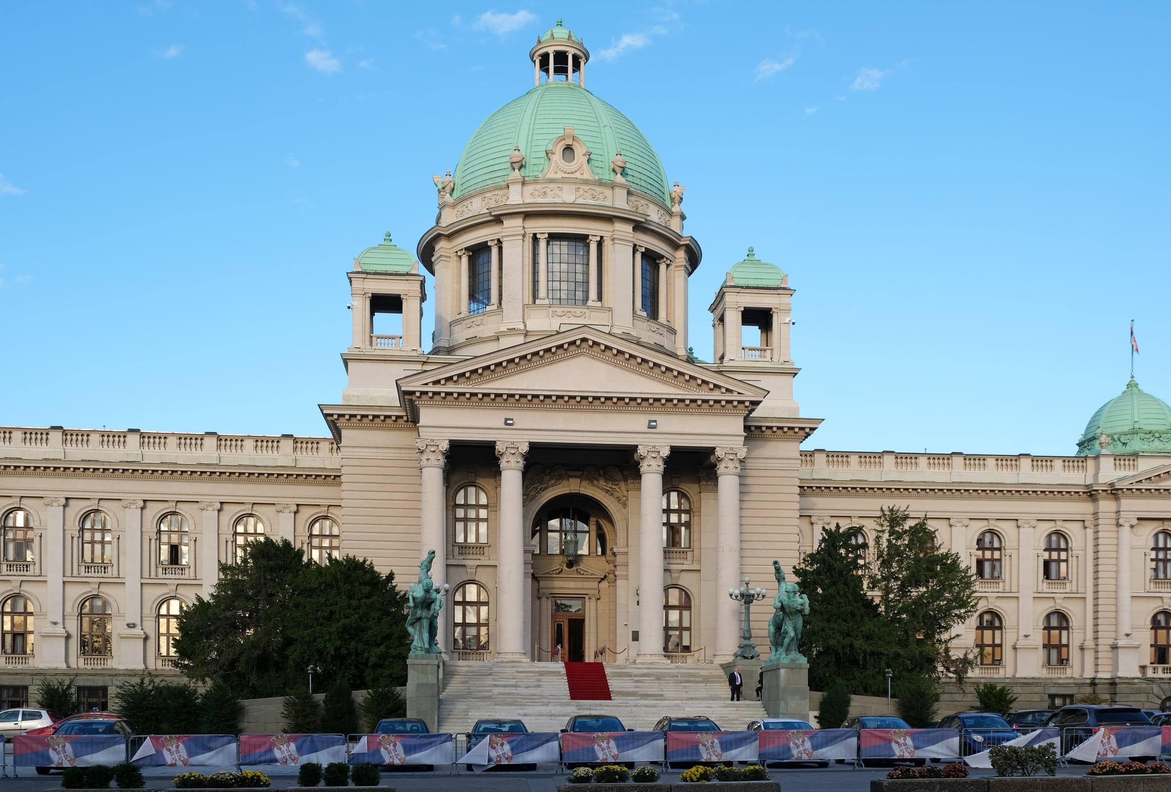 The National Assembly of Serbia in Belgrade seen head-on: a grand neoclassical building with a green copper dome, a columned portico over a red-carpeted staircase, and Serbian-flag banners along the barriers out front.