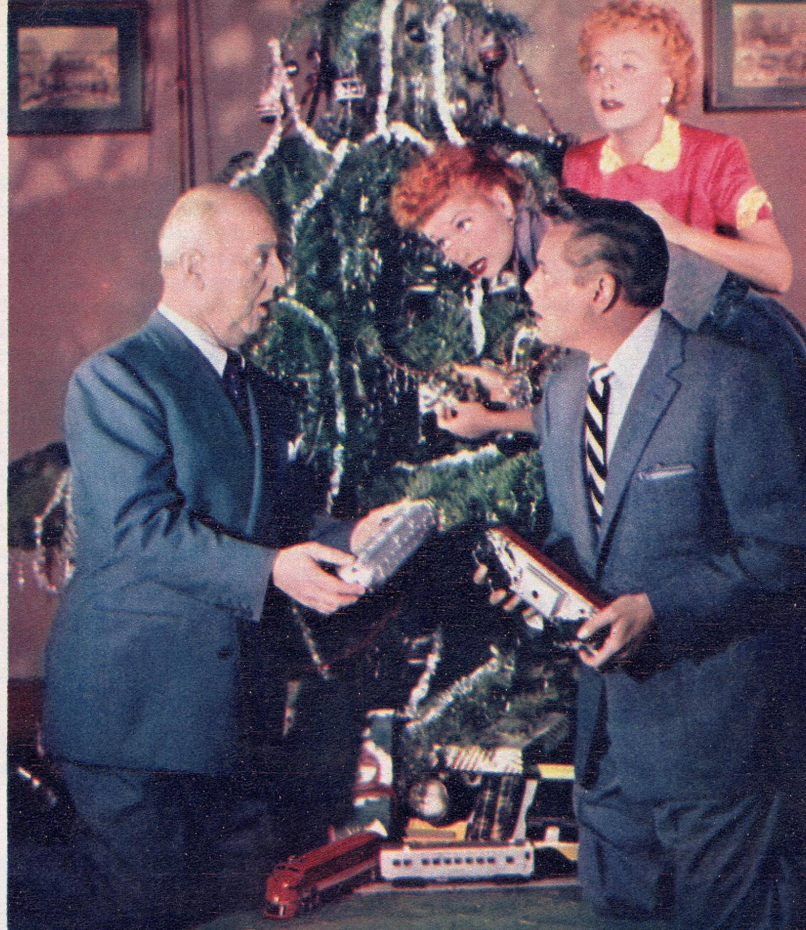 William Frawley and Desi Arnaz hold toy trains beside a decorated Christmas tree as Lucille Ball peers out from the branches and Vivian Vance looks on from behind.