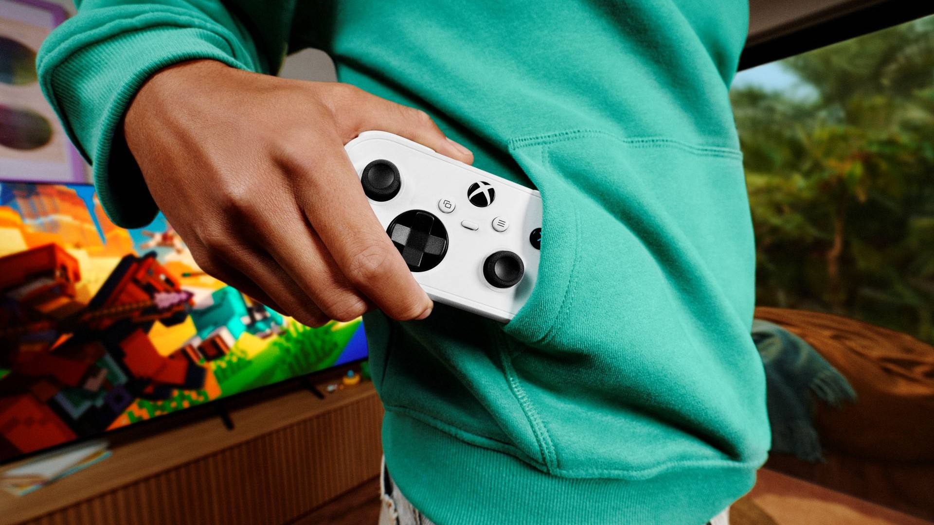 A hand slides a small white Xbox Nano Controller into the front pocket of a teal hoodie, with a Minecraft-style game on a TV in the background.