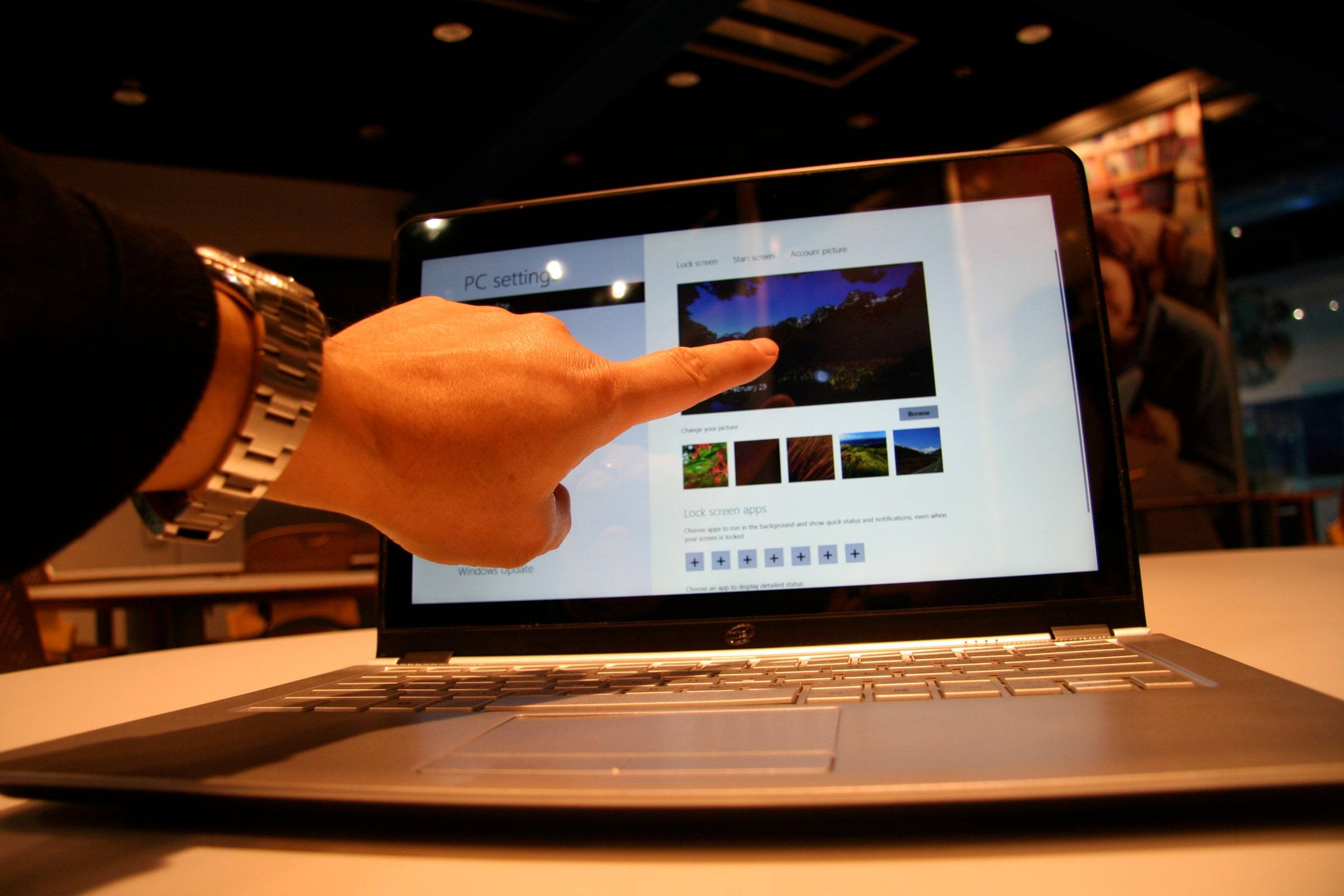 A person's hand reaches toward the touchscreen of an open silver laptop displaying a settings screen, at a brightly lit technology demonstration counter.