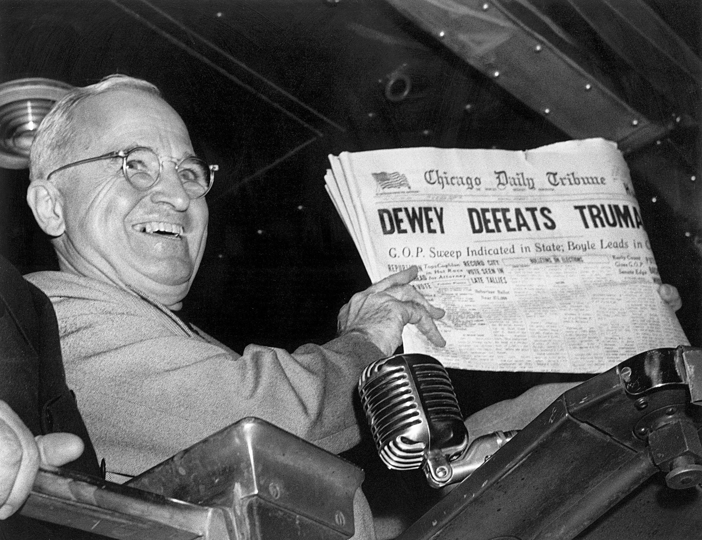 A black-and-white photo of a grinning Harry Truman in glasses holding up a Chicago Daily Tribune front page with the headline DEWEY DEFEATS TRUMAN, a vintage microphone in front of him.