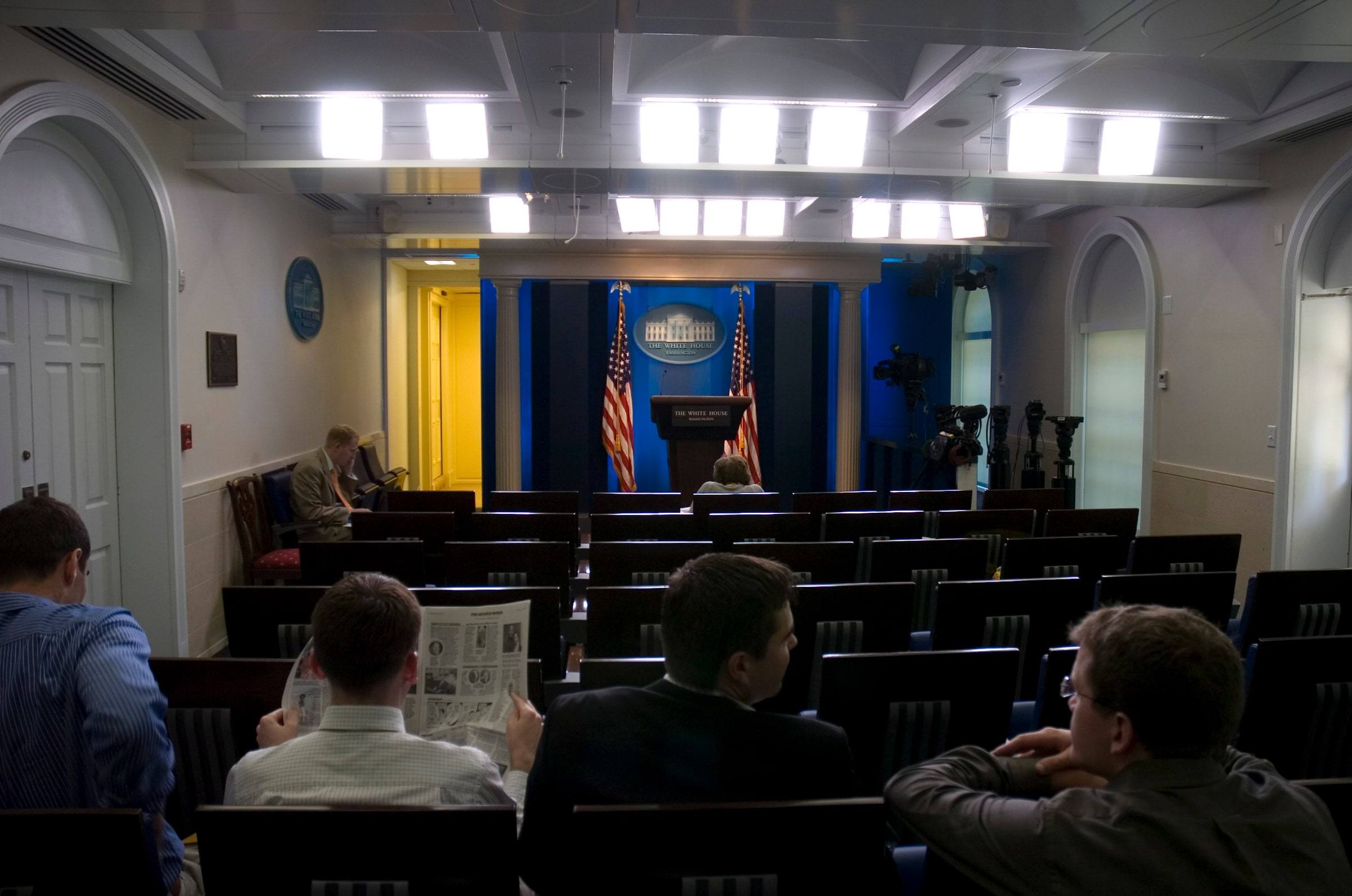 The White House press briefing room seen from the back, rows of seats facing an empty podium with two flags, a few reporters waiting in the seats.